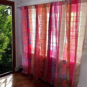 Può includere: Un set di tende trasparenti colorate con un design a patchwork in tonalità rosa, arancio e marrone. Le tende sono appese in una stanza con una finestra che si affaccia su un lussureggiante albero verde.