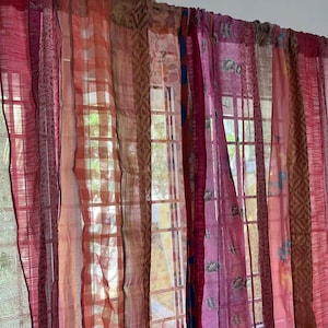 May include: A set of sheer curtains in various shades of pink and red, with different patterns, including stripes, checks, and floral designs. The curtains are hung on a window with a view of a green, leafy background.