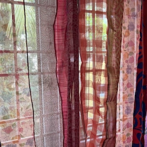 May include: A set of sheer curtains in various shades of red, pink, and white with different patterns. The curtains are hanging in a window and are letting in light.