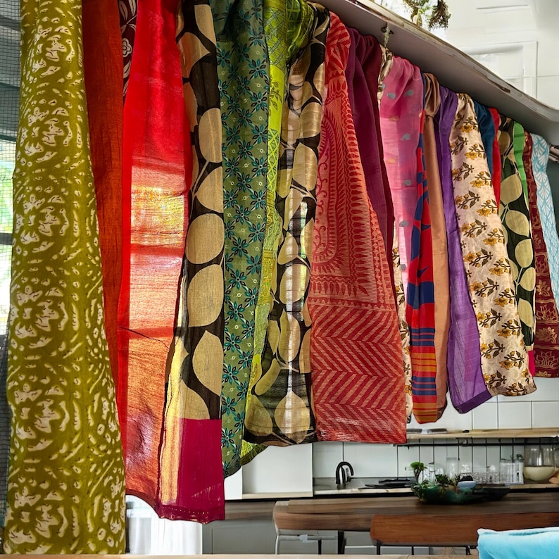 May include: A collection of colorful patterned fabric panels hanging from a ceiling rail. The panels are made of silk and have a variety of designs, including floral, geometric, and abstract patterns. The panels are a mix of bright colors, including red, orange, yellow, green, blue, and purple.