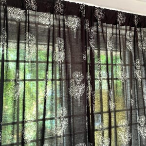 May include: Dark brown sheer curtains with a white floral pattern. The curtains are hanging in front of a window, allowing natural light to filter through. The floral design is a repeating pattern of stylized flowers and leaves.