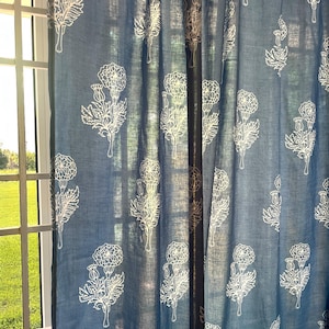 May include: Blue floral curtains with a white flower and leaf pattern. The curtains are hanging in front of a window, with sunlight shining through. The fabric appears to be a lightweight linen or cotton.