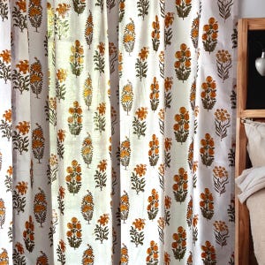 May include: A white curtain panel featuring a repeating block-printed pattern of orange and yellow flowers with green stems and leaves. The fabric is semi-sheer, allowing light to pass through, and hangs in vertical folds with a consistent floral motif.