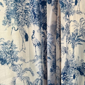 Op de afbeelding: Een stoffen gordijn met een blauw-wit toile-patroon. Het ontwerp toont gedetailleerde illustraties van hoge tropische palmbomen, weelderig gebladerte en diverse vogels, waaronder kraanvogels, die in het dichte botanische landschap staan afgebeeld.