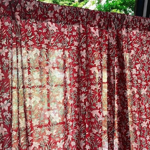 May include: A red fabric curtain featuring a repeating floral pattern in light pink and muted green. The textile has a semi-sheer quality, allowing light to pass through the fabric, highlighting the intricate botanical print and gathered top.