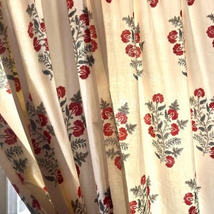May include: A cream-colored curtain featuring a repeating block-print pattern of red flowers with dark gray stems and leaves. The fabric is gathered into vertical folds, creating a textured appearance with light and shadow across the surface.