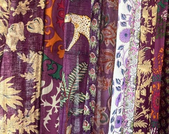 Könnte beinhalten: Eine Sammlung von Stoffbahnen mit Mustern in Lila, Gold und Weiß. Die Designs umfassen Blumen-, Paisley- und Tierdrucke, wie einen Leoparden. Die Stoffe sind vertikal angeordnet und erzeugen eine visuell reiche Darstellung von Texturen und Farben.
