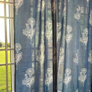 May include: A blue curtain panel featuring a repeating white line-art floral pattern of thistle-like blossoms and leaves. The fabric has a visible weave texture and is hung in vertical folds, allowing light to pass through the semi-sheer material.