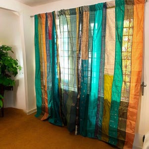 May include: A set of teal, green, gold, and orange patchwork curtains made from recycled saris. The curtains are hanging in a window and are made of various fabrics.