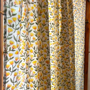May include: Curtains with a repeating floral pattern. The design features yellow flowers with orange centers and green leaves on a white background. The curtains are hanging in front of a window.