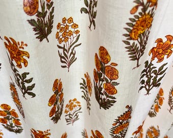 Puede incluir: Una cortina de tela blanca con un patrón repetitivo de estampado en bloque de flores naranjas y amarillas con tallos y hojas verdes. El tejido tiene una textura visible y está drapeado en suaves pliegues verticales, mostrando varios motivos florales.