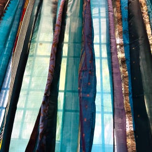 Pode incluir: Uma janela com vista para o exterior é coberta por uma cortina feita de várias tiras de tecido em vários tons de azul, roxo e dourado. O tecido é transparente e permite que a luz brilhe.