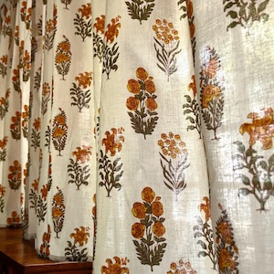 Peut inclure: Rideaux blanc cassé avec un motif imprimé répétitif de fleurs stylisées dans des tons jaune moutarde et vert olive. Le tissu présente une texture tissée visible et tombe en plis verticaux, affichant de multiples motifs floraux.
