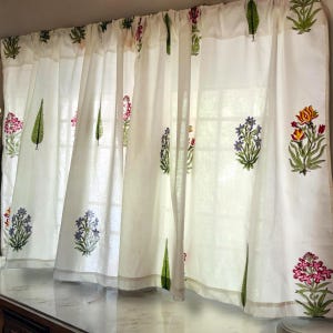 Puede incluir: Una cortina blanca con un patrón repetitivo de bloques impresos de diversos motivos florales y hojas verdes. El diseño incluye pequeños grupos de flores rosas, azules, amarillas y rojas distribuidas por la tela, que cuelga en pliegues suaves.