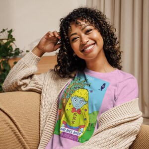Op de afbeelding: Een roze T-shirt met een cartoonillustratie van een meisje met geel haar en een blauw gezicht. De illustratie is omgeven door een blauwe rand en bevat de tekst "Baby Bee's Boot" en "Eiren".