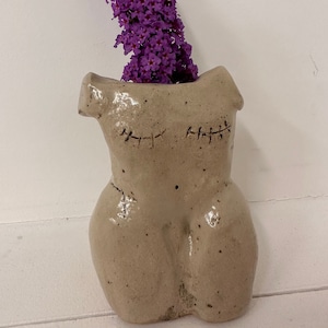 Puede incluir: Un jarrón de cerámica con forma de torso con detalles cosidos, que contiene un ramo de flores moradas.