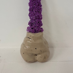 May include: A ceramic vase shaped like a human body with a cluster of purple flowers in the top opening.