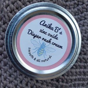 May include: A round, silver-colored tin of Anika B's zinc oxide diaper rash cream. The label features a pink circle with the product name and a blue and yellow bee illustration. The words "smooth & all natural" are also on the label.