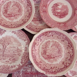 May include: A collection of white ceramic plates featuring intricate pink transferware patterns depicting rural landscapes, cottages, and trees. The plates have scalloped or smooth edges and are decorated with detailed floral and leaf borders.