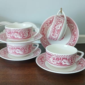 May include: A set of three cream-colored ceramic teacups and saucers featuring a detailed red transferware pattern of pastoral landscapes and buildings. The cups have curved handles and the saucers have a matching decorative border design.