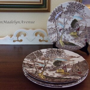 May include: A set of ceramic plates featuring a brown and white transferware scene of a village with trees, buildings, and a path. One plate is displayed on a black stand, while others are stacked below. The text OnMadelynAvenue is visible.