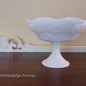 May include: A white milk glass pedestal bowl featuring a raised relief pattern of grape clusters and leaves around the exterior. The bowl has a scalloped rim and sits on a circular base. The text OnMadelynAvenue is visible in the lower left corner.