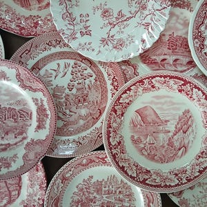 May include: A collection of ceramic plates featuring intricate red and white transferware patterns. Each plate displays detailed landscape scenes, including bridges, buildings, and coastal views, framed by ornate floral and leaf borders.