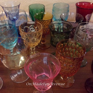 May include: A collection of multi-colored glass goblets featuring various textures, patterns, and stem designs in shades of red, blue, green, amber, and pink, with the text OnMadelynAvenue printed in white at the bottom center of the image.