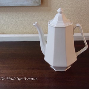 May include: A white ceramic teapot featuring an octagonal body, a matching lid with a pointed finial, a curved spout, and an angular handle. The text OnMadelynAvenue is printed in white in the lower left corner of the image.