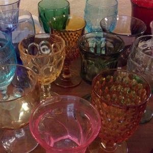 May include: A collection of assorted vintage-style glass goblets in various colors including amber, red, blue, green, and pink. The drinking glasses feature diverse textures such as hobnail, diamond patterns, smooth surfaces, and decorative rims.