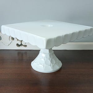 May include: A white square cake stand with a smooth, flat top surface and a scalloped edge design. The pedestal base features a raised grid pattern, providing a stable foundation for the square platform. The entire piece has a glossy finish.