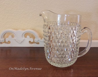 May include: A clear glass pitcher featuring a prominent diamond-point texture across its exterior surface. The vessel includes a curved handle on the right side, a wide pouring spout, and the text OnMadelynAvenue printed in the lower left corner.