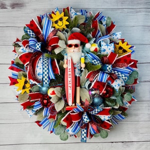 May include: A festive wreath with red, white, and blue ribbons, ornaments, and greenery. A Santa Claus nutcracker figure in sunglasses holds a surfboard that says "Surf's Up!". Decorative suns and beach balls complete the summer holiday theme.
