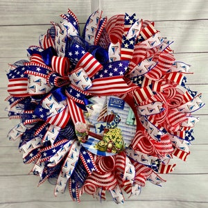 May include: A red, white, and blue patriotic wreath with a lake scene in the center. The wreath is made of mesh and ribbon and features a variety of nautical and patriotic designs. The lake scene features a boat, a dock, and a sign that says "Lake".