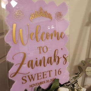Op de afbeelding: Een paars en gouden welkomsbord met de tekst "Welcome to Zainab's Sweet 16 November 30, 2021". Het bord is versierd met een gouden kroon en rozen.