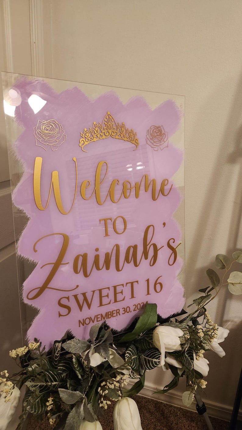 Op de afbeelding: Een paars en gouden welkomsbord met de tekst "Welcome to Zainab's Sweet 16 November 30, 2021". Het bord is versierd met een gouden kroon en rozen.