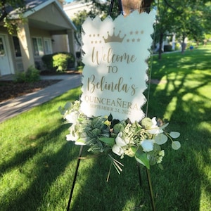 May include: A clear acrylic sign with gold lettering on an easel. The sign reads "Welcome to Belinda's Quincea&ntilde;era September 24, 2022". The sign is decorated with a gold crown and stars.