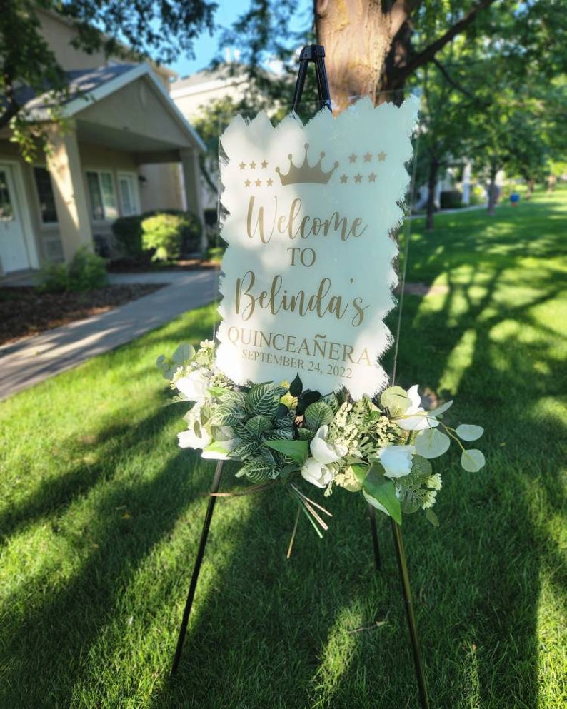 May include: A clear acrylic sign with gold lettering on an easel. The sign reads "Welcome to Belinda's Quincea&ntilde;era September 24, 2022". The sign is decorated with a gold crown and stars.