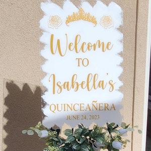 Op de afbeelding: Een wit acryl bord met gouden letters en een gouden kroon en rozen. Het bord zegt "Welkom bij Isabella's Quincea&ntilde;era op 24 juni 2023".