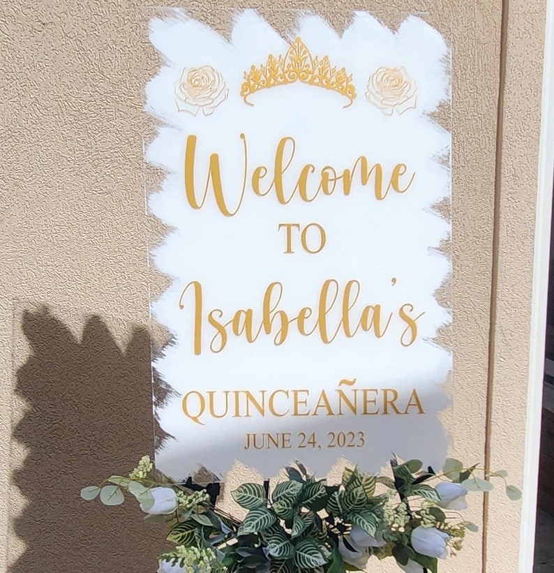 Op de afbeelding: Een wit acryl bord met gouden letters en een gouden kroon en rozen. Het bord zegt "Welkom bij Isabella's Quincea&ntilde;era op 24 juni 2023".