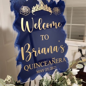 Op de afbeelding: Een blauw en gouden welkomsbord voor een quincea&ntilde;era. Het bord zegt "Welkom bij Briana's Quincea&ntilde;era op 27 mei 2023".