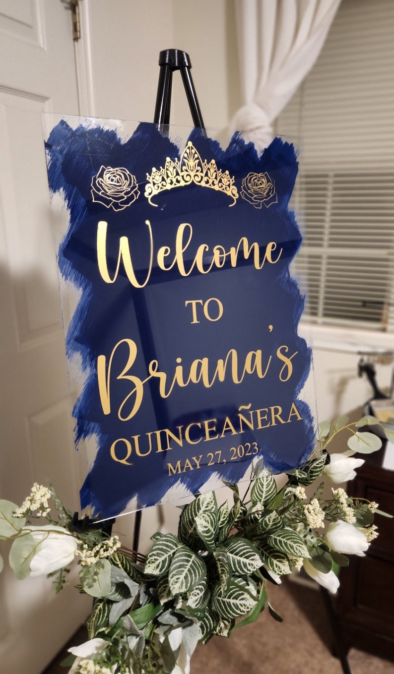 Op de afbeelding: Een blauw en gouden welkomsbord voor een quincea&ntilde;era. Het bord zegt "Welkom bij Briana's Quincea&ntilde;era op 27 mei 2023".