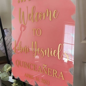 May include: A pink acrylic welcome sign with gold lettering and a gold crown. The sign reads "Welcome to [Name]'s Quincea&ntilde;era April 16, 2023".