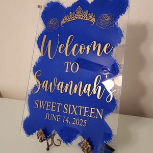 Op de afbeelding: Een blauw en gouden welkomsbord voor een Sweet Sixteen feest. Het bord zegt "Welkom bij Savannah's Sweet Sixteen June 14, 2025".