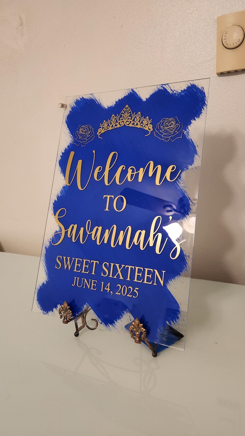 Op de afbeelding: Een blauw en gouden welkomsbord voor een Sweet Sixteen feest. Het bord zegt "Welkom bij Savannah's Sweet Sixteen June 14, 2025".