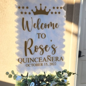 May include: A white sign with gold lettering that says "Welcome to Rose's Quincea&ntilde;era October 15, 2022". The sign has a gold crown and stars at the top.