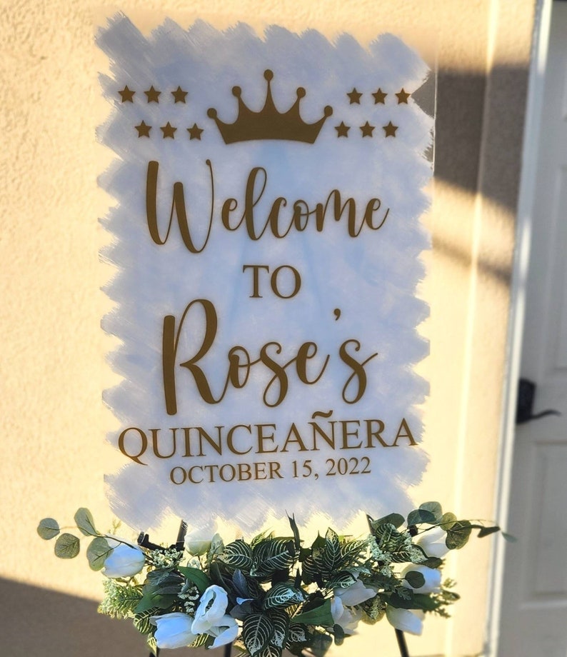 May include: A white sign with gold lettering that says "Welcome to Rose's Quincea&ntilde;era October 15, 2022". The sign has a gold crown and stars at the top.