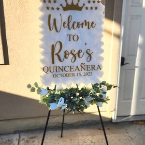 May include: White welcome sign with gold lettering that reads "Welcome to Rose's Quincea&ntilde;era October 15, 2022". The sign is decorated with a gold crown and stars. The sign is on a black easel with white flowers.
