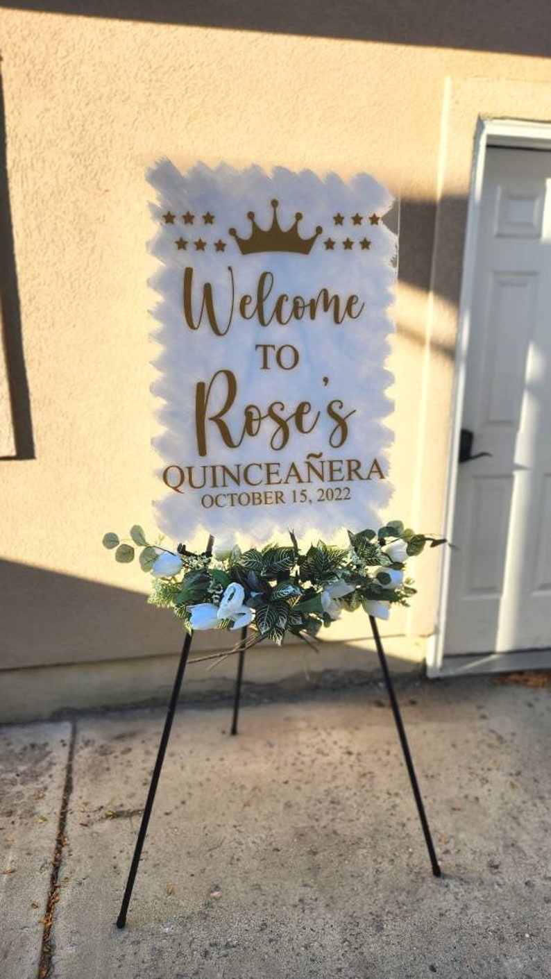May include: White welcome sign with gold lettering that reads "Welcome to Rose's Quincea&ntilde;era October 15, 2022". The sign is decorated with a gold crown and stars. The sign is on a black easel with white flowers.