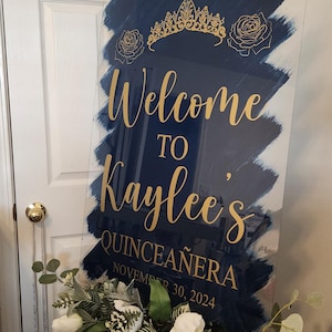 Op de afbeelding: Een blauw en gouden welkomsbord voor een Quincea&ntilde;era. Het bord zegt "Welkom bij Kaylee's Quincea&ntilde;era op 30 november 2024".